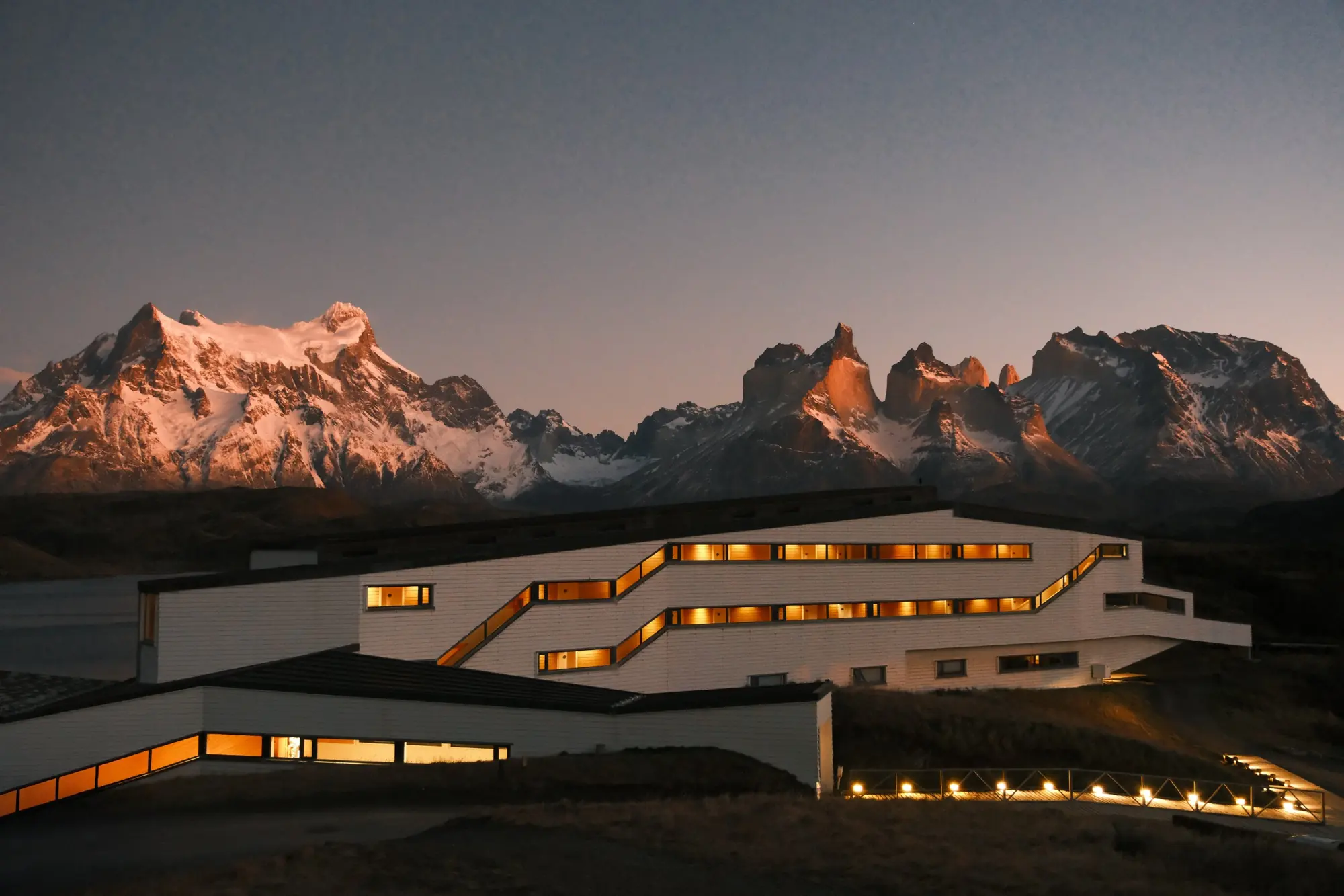 Explora Patagonia's long white lodge, its windows lit at dusk, below Paine Grande and the Cuernos del Paine